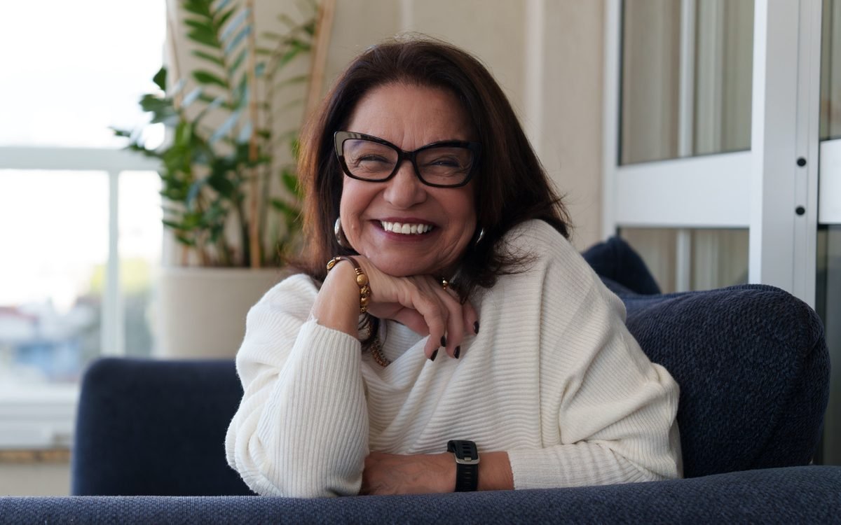 Elsa Saldanha sentada em um sofá, rindo, com a luz da janela ao fundo