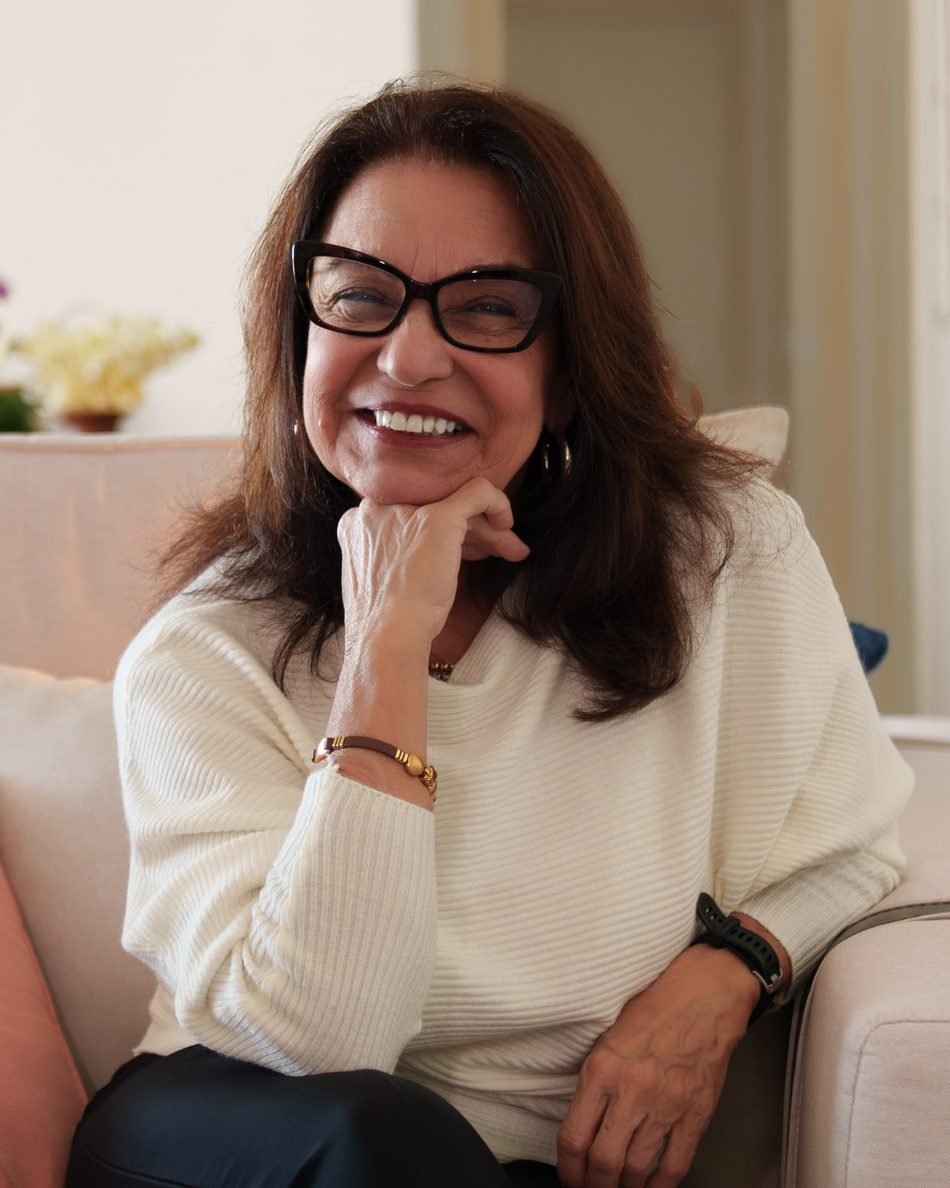 Elsa Saldanha sentada em uma poltrona clara, sorrindo, com a mão apoiada no queixo